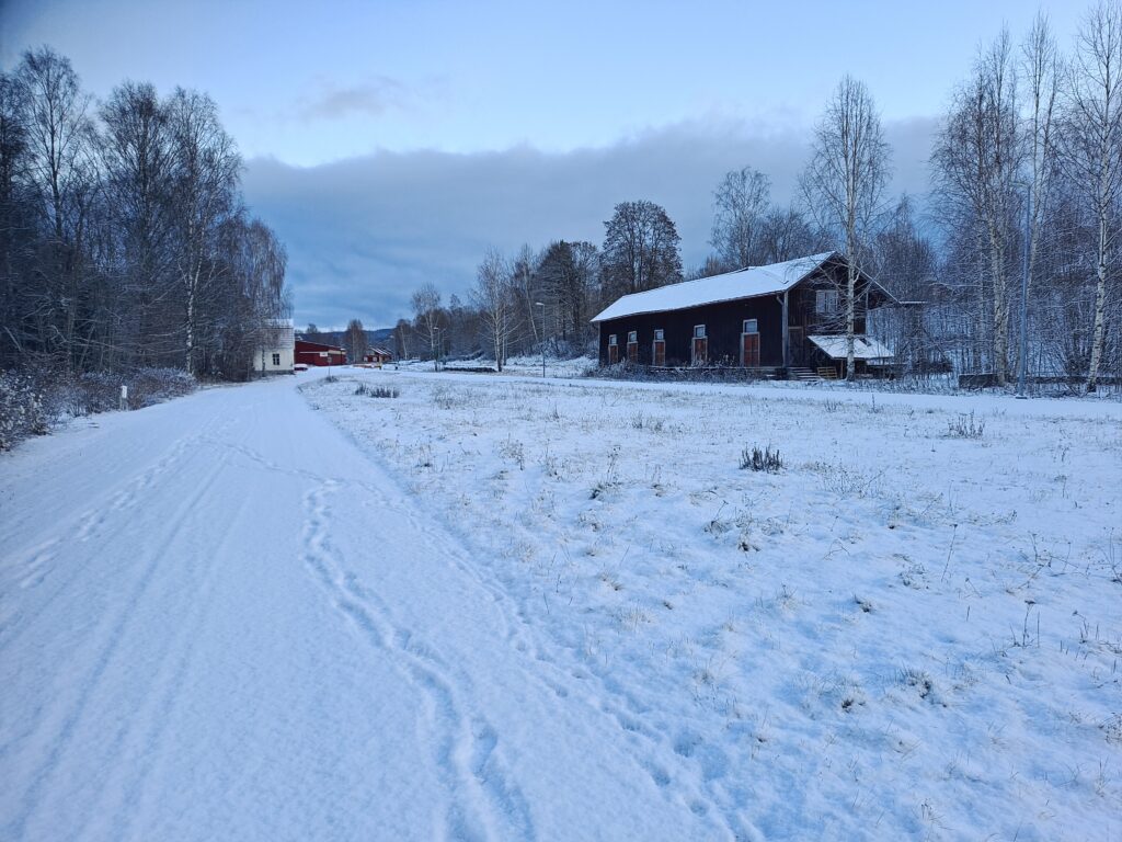 A snow-covered road in Sweden; the ground is lighter than the sky. On the right, a dilapidated structure, originally used to load goods onto the long-gone railway track.